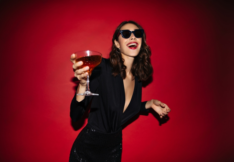 smiling woman in black dress and sunglasses holds red cocktail near red wall